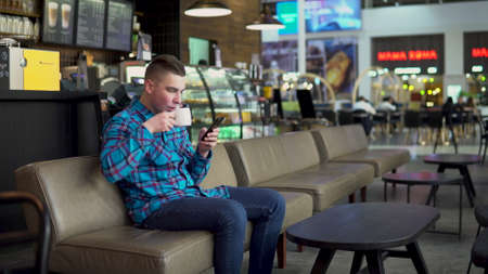 A young man is sitting in a cafe with a phone. The man is in correspondence on a smartphone and drinks coffee. Close-up.の写真素材