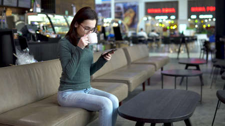 A young woman is sitting in a cafe with a phone. The girl is in correspondence on a smartphone and drinks coffee. Close-up.の写真素材