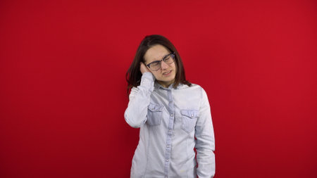 A young woman with glasses has an ear ache and she holds it in her hands. Shooting on a red background.の写真素材
