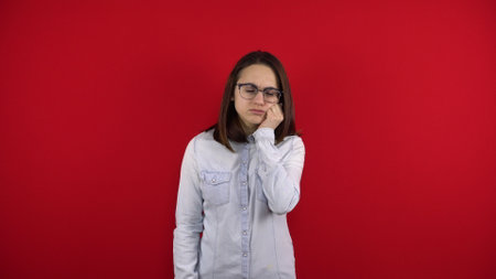 A young woman wearing glasses has a toothache and touches her cheek with her hand. Shooting on a red background.の写真素材