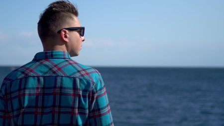 A young man looks at the sea. A man in a blue shirt admires nature. A man stands with his back to...