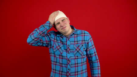 The young man has a bandage on his head, he is holding it with his hands because of the pain. Shooting on a red background.の写真素材