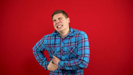 A young man has a liver ache, he holds his side with his hand. Shooting on a red background.の写真素材