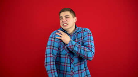 A young man has a sore shoulder and he touches it with his hand. Shooting on a red background.の写真素材