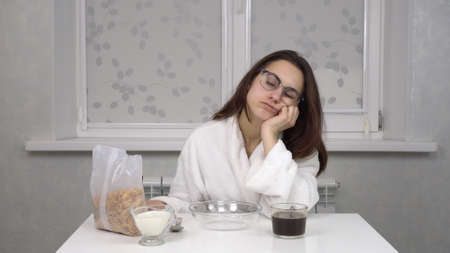 Sleepy young woman in a white robe eats cereals and drinks coffee. Young girl with glasses at home in the morning kitchen.の写真素材