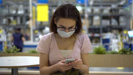A young Asian girl in glasses and a mask sits in a store with a phone in her hands.の写真素材
