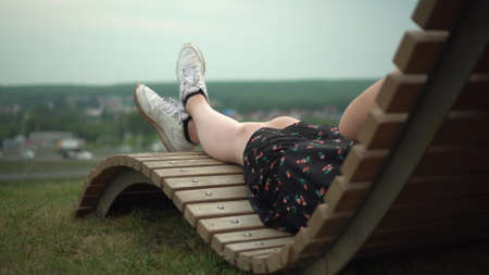 A young girl in a dress and sneakers lies on a deck chair on the grass. Girls legs close up.