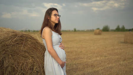 A young pregnant woman stands near a haystack in a field. A girl with glasses and a blue dress.の写真素材