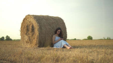 A young woman sits near a haystack in a field with a phone in her hands. A girl with glasses and a blue dress is texting on the phone.の写真素材