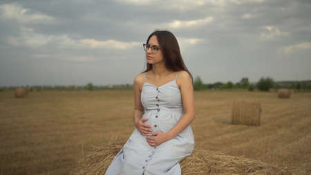 Young pregnant woman sits on a haystack in a field. Girl with glasses and a blue dress.の写真素材