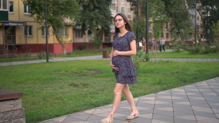 A young pregnant girl walks along the alley and touches her stomach with her hands. Girl with glasses and a dress in the park.の写真素材