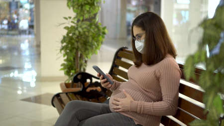 A young pregnant woman in glasses and a medical mask sits with a phone on a bench. Girl in the mall...