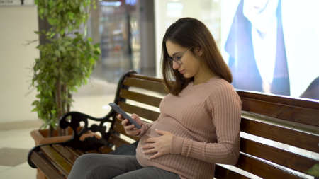 A young pregnant woman in glasses sits with a phone on a bench. Girl in the mall and uses a smartphone.の写真素材