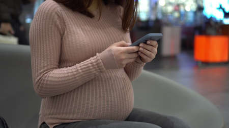 A young pregnant woman in glasses sits with a phone on a bench. Girl in the mall and uses a smartphone. Closeup.の写真素材