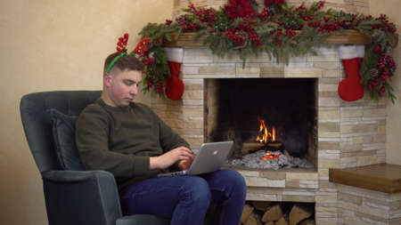 A young man sits in an armchair by the fireplace with horns on his head and is typing on a laptop. A man writes an email to Santa Claus.の写真素材