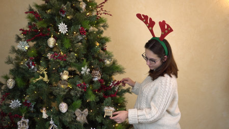 A young pregnant woman in glasses and a sweater with horns on her head examines the toys on the Christmas tree. Christmas mood.の写真素材
