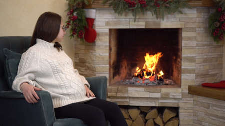 A young pregnant woman with glasses in a sweater sits in an armchair by the fireplace and looks at the fire. Christmas mood.の写真素材