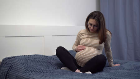 Young pregnant woman with glasses stroking her belly. The girl in the bedroom on the bed.の写真素材