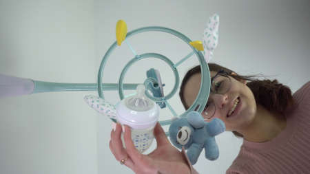 A young mother gives a bottle of milk to a newborn baby lying in a cradle with a baby mobile with toys. First-person view from the bottom up.の写真素材