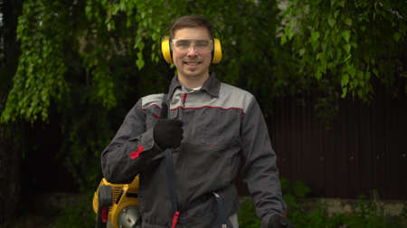 A young man from the special services mows the lawn with a petrol trimmer. A man in glasses and headphones looks at the camera, smiles and shows a thumbs up.の写真素材