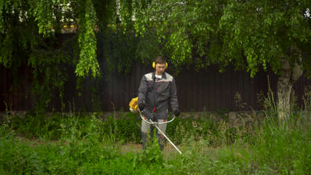A young man from the special services mows the lawn with a petrol trimmer. A man in glasses and...