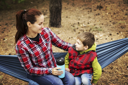 Camping in the forest. Mom and son are sitting in a hammock and looking at the fire.の写真素材