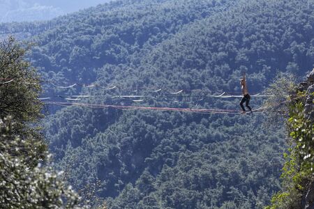 A man is walking along a stretched sling. Highline in the mountains. Man catches balance. Performance of a tightrope walker in nature. Highliner on the background of valley.の写真素材