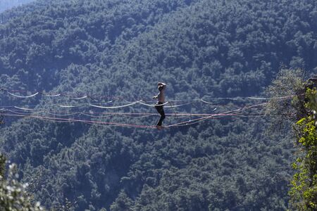A man is walking along a stretched sling. Highline in the mountains. Man catches balance. Performance of a tightrope walker in nature. Highliner on the background of valley.の写真素材