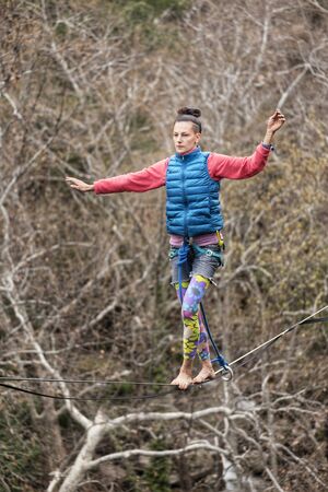 A woman is walking along a stretched sling. Highline in the mountains. Woman catches balance. Performance of a tightrope walker in nature. Highliner on the background of valley.の写真素材