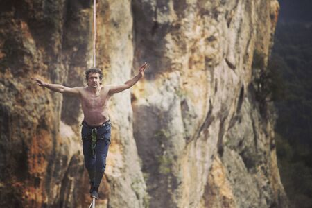 A man is walking along a stretched sling. Highline in the mountains. Man catches balance. Performance of a tightrope walker in nature. Highliner on the background of the mountains.の写真素材