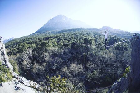 A man is walking along a stretched sling. Highline in the mountains. Man catches balance. Performance of a tightrope walker in nature. Highliner on the background of the mountains.の写真素材