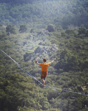 A man is walking along a stretched sling. Highline in the mountains. Man catches balance. Performance of a tightrope walker in nature. Highliner on the background of the mountains.の写真素材