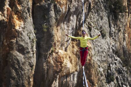 A woman is walking along a stretched sling. Highline in the mountains. Woman catches balance. Performance of a tightrope walker in nature.の写真素材