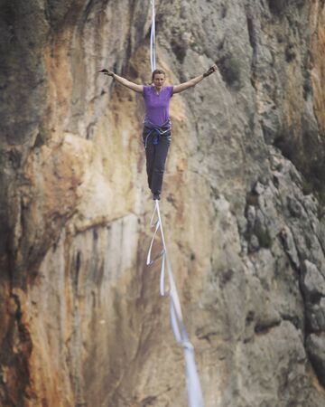A woman is walking along a stretched sling. Highline in the mountains. Woman catches balance. Performance of a tightrope walker in nature.の写真素材