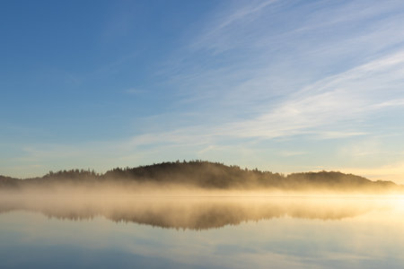 Golden sunrise over calm autumn sea with forest on the shore and gentle fog above the water, peaceful natural seascape and morning atmosphere.の写真素材