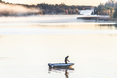 Fisherman on a small boat fishing at sunrise in calm foggy sea, forest coastline visible in the distance, tranquil autumn seascape with warm morning light.の写真素材