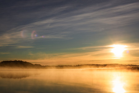 Golden sunrise over calm autumn sea with forest on the shore and gentle fog above the water, peaceful natural seascape and morning atmosphere.の写真素材