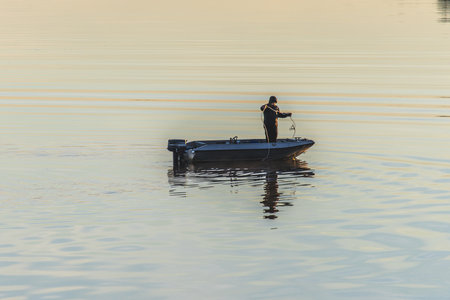 Fisherman on a small boat fishing at sunrise in calm foggy sea, coastline visible in the distance, tranquil autumn seascape with warm morning light.の写真素材