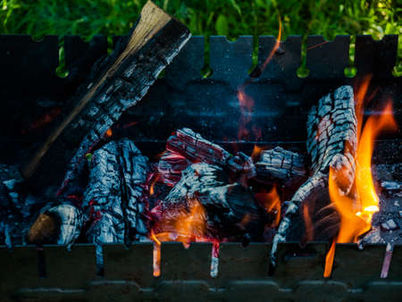 barbecue woods, flame fire, ash coals burning in metal grill preparing for BBQ outdoors green grass in sunny daytime backyard. going smoke up from burning wooden logs, campfire orange flamesの写真素材