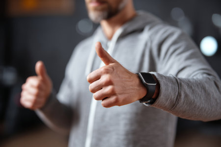 A man wearing a gray hoodie stands indoors, smiling while giving a thumbs up with both hands. He is engaged in a fitness routine, showcasing his smartwatch on his wrist.の素材