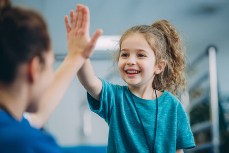 A young girl with curly hair smiles brightly as she gives a high five to an adult during an engaging activity in a lively indoor setting. The atmosphere feels warm and supportive.の素材