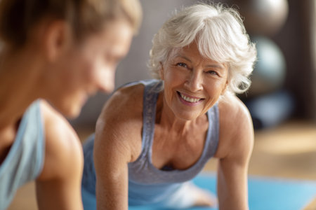 In a bright yoga studio, a senior woman smiles while practicing yoga alongside a younger instructor. The warm atmosphere supports a sense of community and well-being.の素材