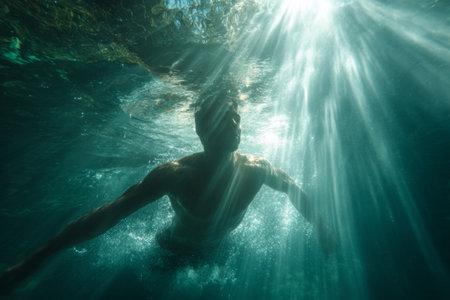 A man swims gracefully below the surface of the water, illuminated by rays of sunlight filtering through the clear ocean. The tranquil scene captures the beauty of nature.の素材