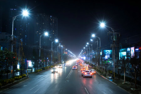 Cars drive along a wide city street illuminated by bright streetlights and colorful signs. The scene captures the energy of urban life during the night in a bustling area.の素材