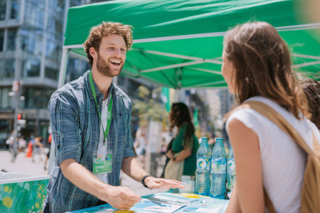 A cheerful young man is talking to a visitor at a green event booth set up in a lively city street. Sunlight highlights the atmosphere of engagement and community interaction.の素材