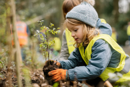 Children actively participate in a community tree planting event, wearing bright vests. They carefully plant small trees in the ground, contributing to environmental care.の素材