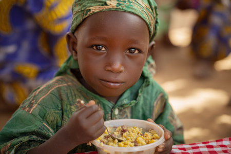 A young child sits at a table in a colorful outdoor setting, happily eating a bowl of corn with meat. The warm sunlight highlights the bright clothing around them.の素材