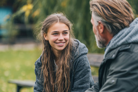 A teenager with long hair smiles while sitting on a bench in a park. An older man with gray hair listens attentively. Yellow leaves surround them as they share a moment.の素材