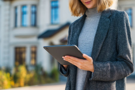 A young woman with wavy hair stands outside a stylish building, focused on a tablet. She wears a gray blazer and a light sweater as she interacts with the device in the bright sun.の素材