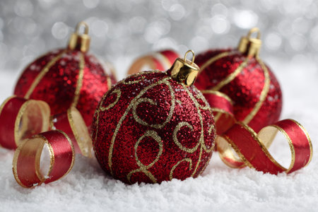 Sparkling red Christmas ornaments with golden patterns are arranged around shiny curling ribbons. The snowy background adds a festive touch, perfect for holiday decorations.の素材